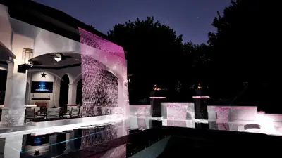 Rain curtain lit magenta falling from the loggia roof beside the pool, fire bowl on the wall